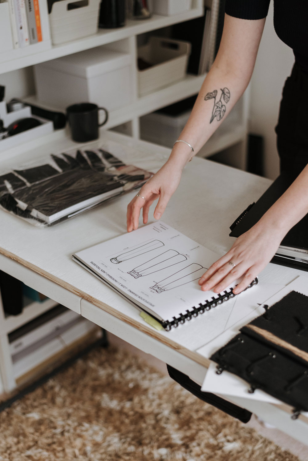 Close-up of a woman's hands at a desk looking through a sketchbook filled with fashion designs for pants.